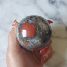 Load image into Gallery viewer, A hand holding a small gray and red African Bloodstone sphere with a wooden stand.
